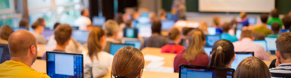 La imagen muestra una clase magistral en la que los estudiantes que ocupan los diversos asientos, hacen uso de sus ordenadores personales para seguir la explicación